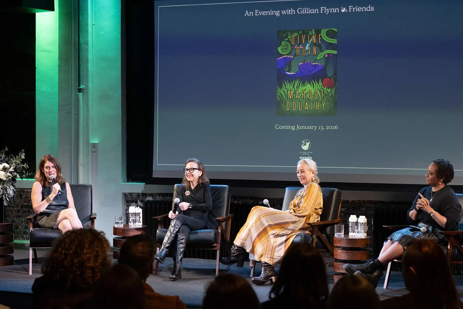 The host and panelist smile as they are talking with the audience. The book cover of Divine Ruin by Margot Douaihy is projected on the screen behind with the text coming January 13th 2026.