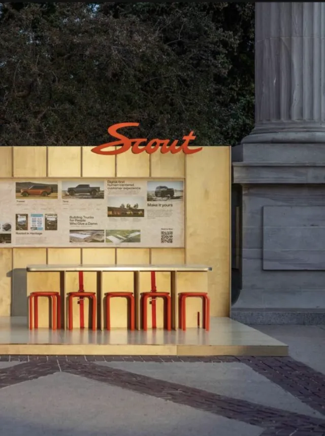 Wooden structure with Scout logomark in upper right and small stools at a bar like counter. Images of the Scout Motors vehicles displayed on the wall of the structure.